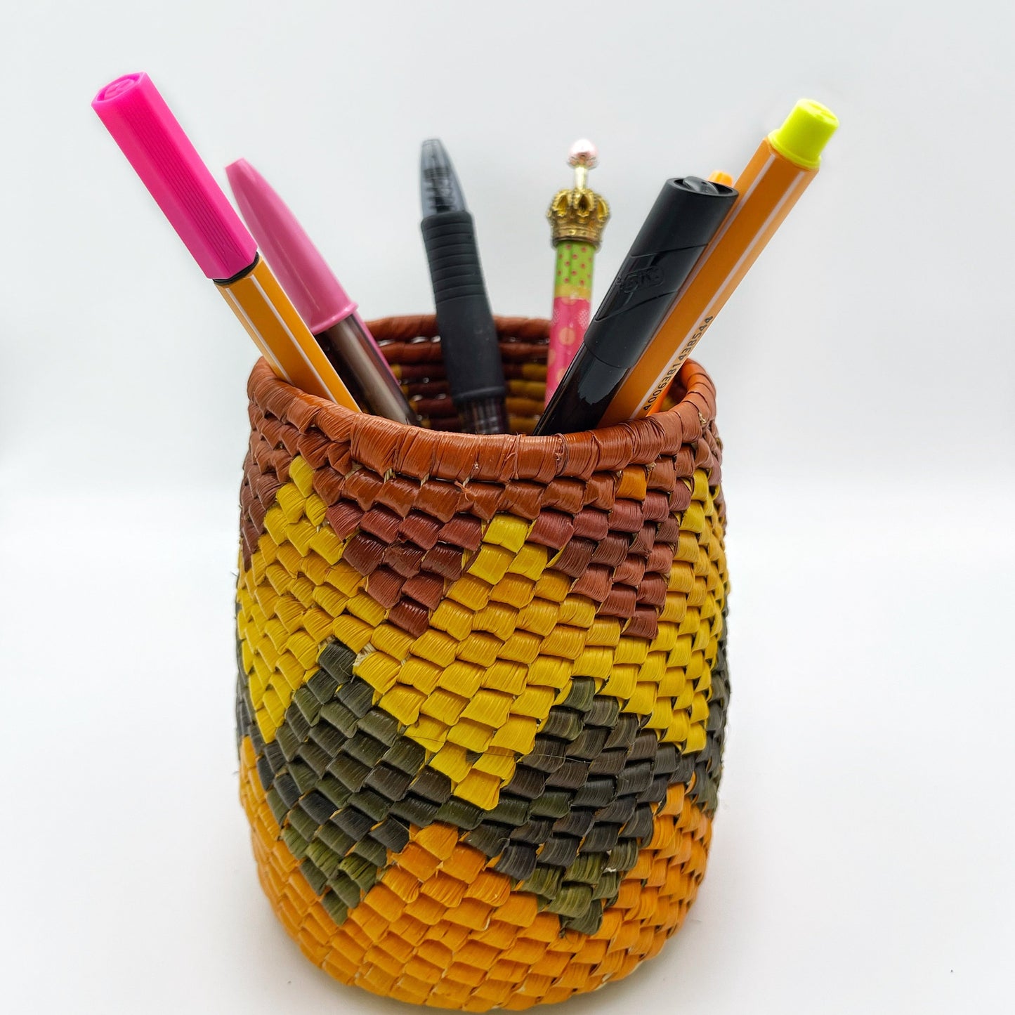 Colorful woven pen holder in yellow, olive, and brown hues, filled with vibrant pens, against a white backdrop.