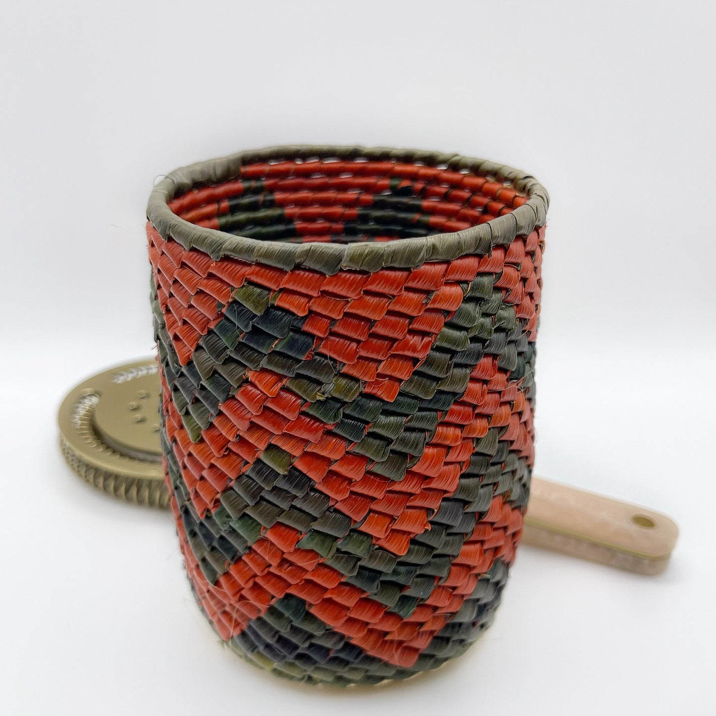 A small woven holder with a checkered pattern in olive green, red, and black, displaying fine craftsmanship.