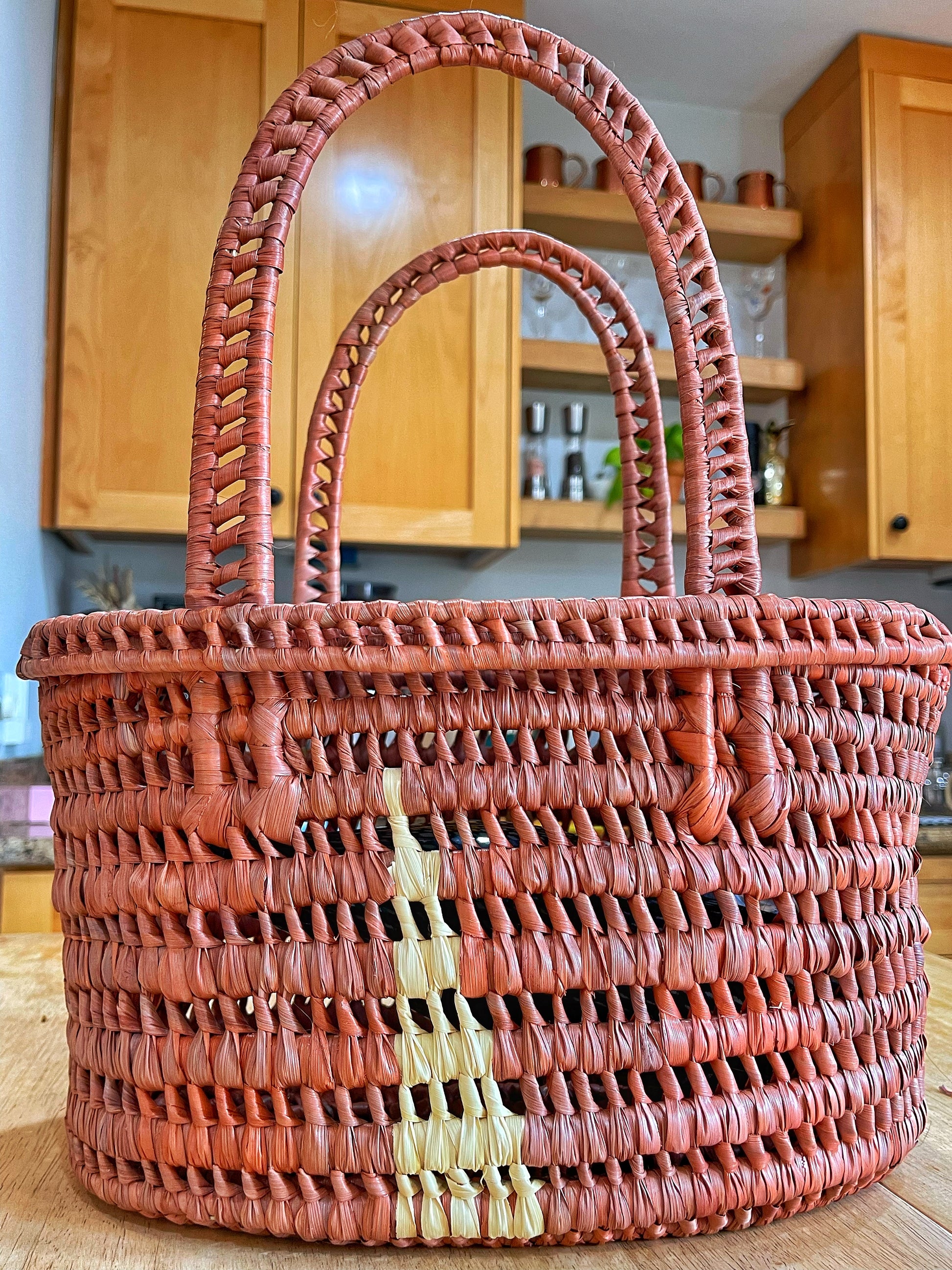 A terracotta-colored woven basket stands against a kitchen backdrop, its craftsmanship highlighted by natural light, with a subtle pattern of cream.