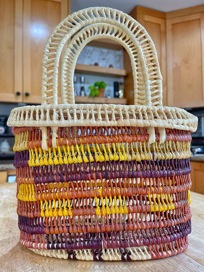 Crafted Tucuma palm basket with alternating bands of yellow, orange, and brown, featuring a tight weave pattern and a robust, spiraled handle in a natural cream hue