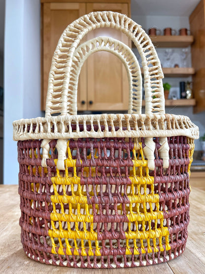 Durable handwoven basket made from Tucuma palm fibers, with cream edges and handles, and a body of yellow, orange, and brown hues in horizontal stripes.