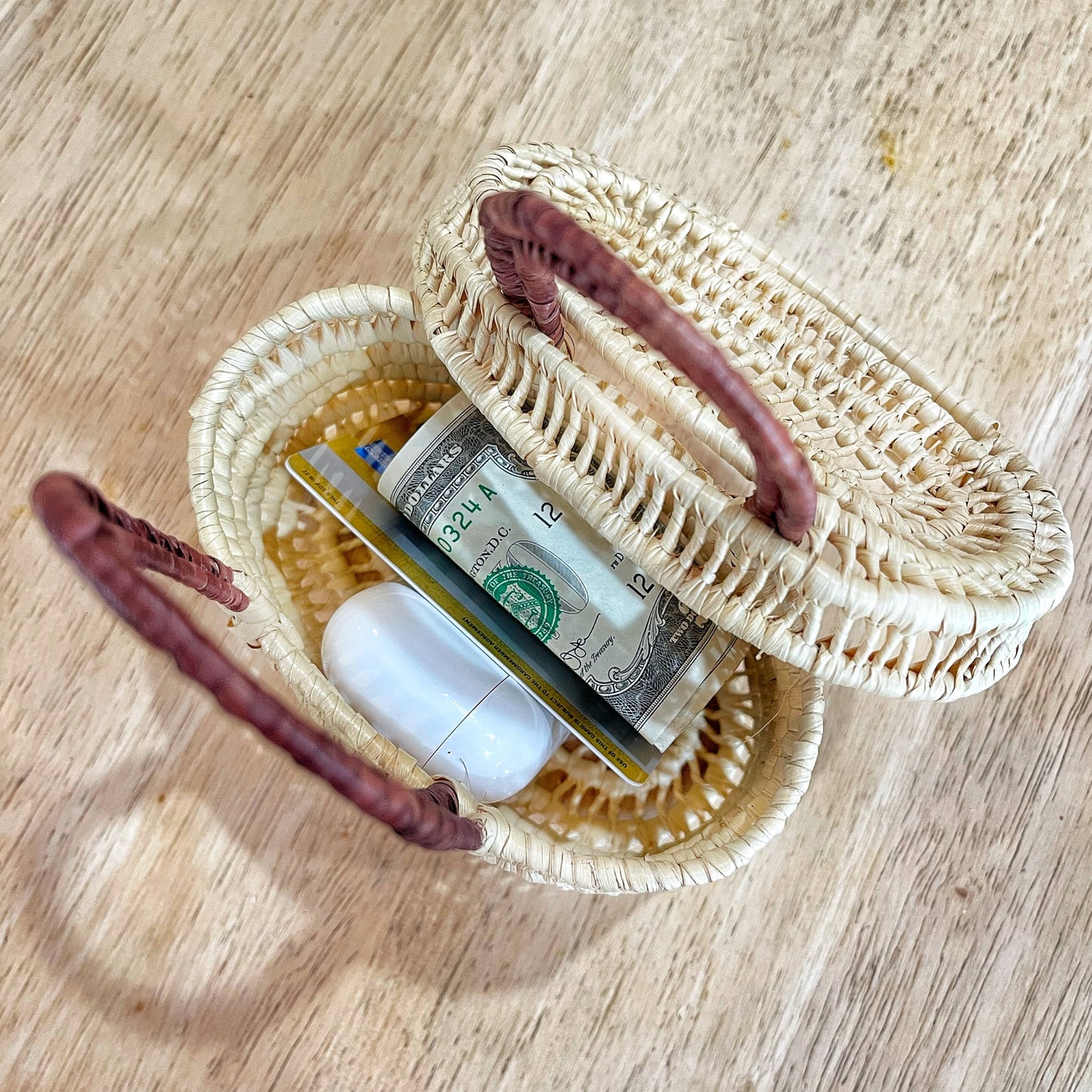 Small, handwoven Amazonian basket with dark handles holds earbuds, cash, cards on a wooden table. Perfect for personal belongings.