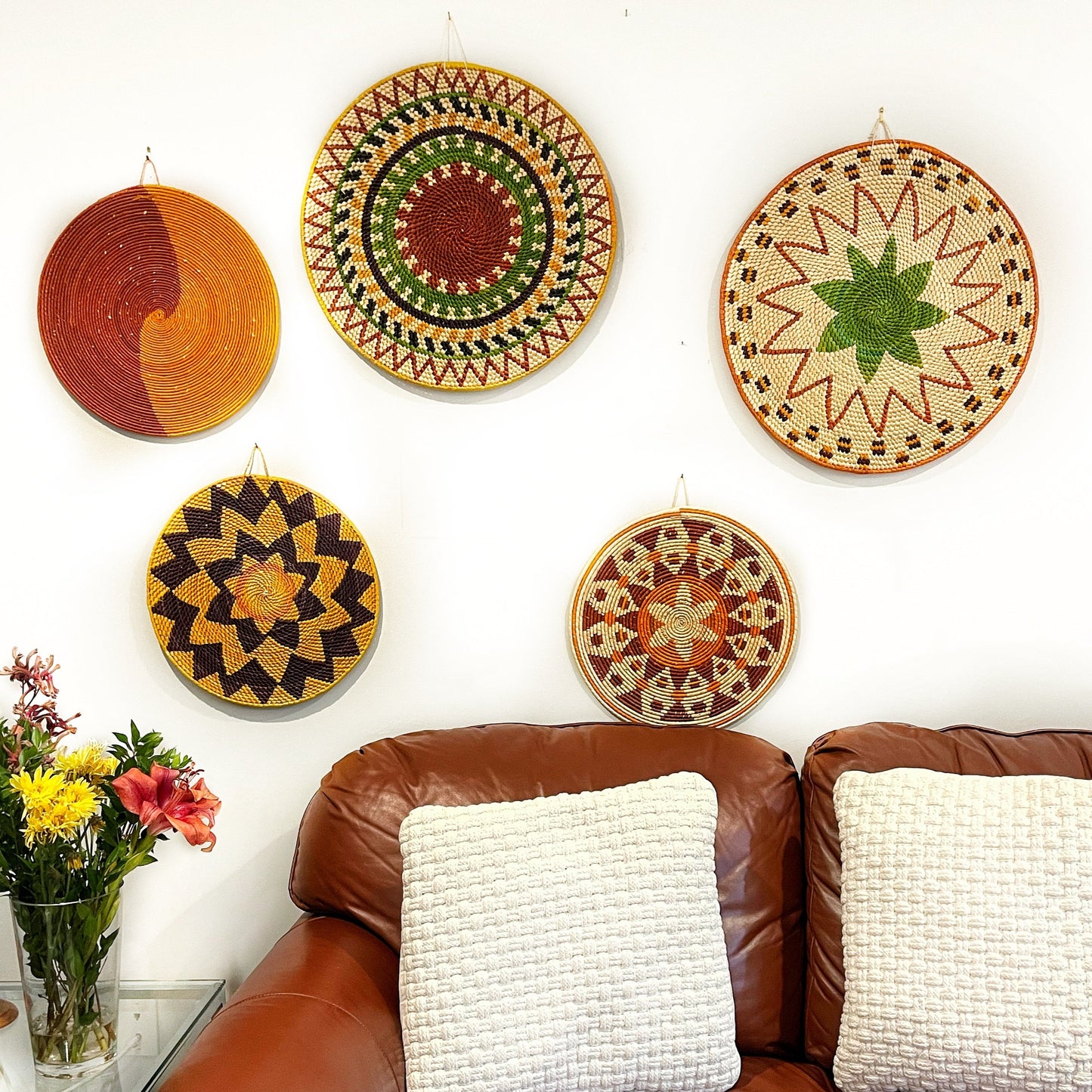 The image features a cozy living room adorned with five distinct mandala wall art pieces, each boasting unique geometric and spiral designs in warm tones of red, yellow, green, and cream, against a white wall above a brown leather couch.