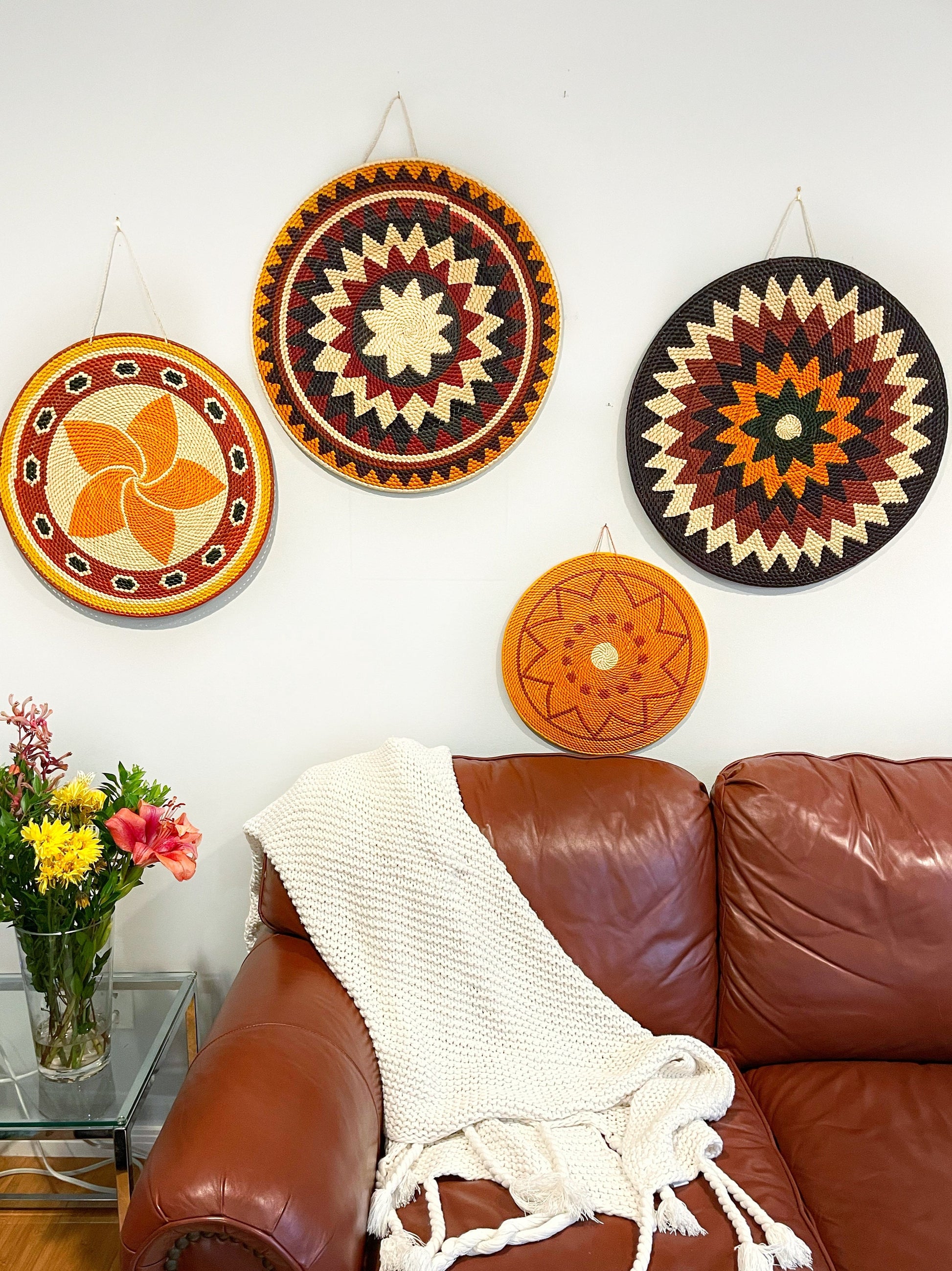 A living room wall adorned with four Amazonian handwoven mandala artworks, each with distinct geometric patterns in warm tones. Below, a brown leather couch is draped with a white knitted throw, complemented by a vase of fresh flowers, infusing the space with cultural richness and artisanal charm.