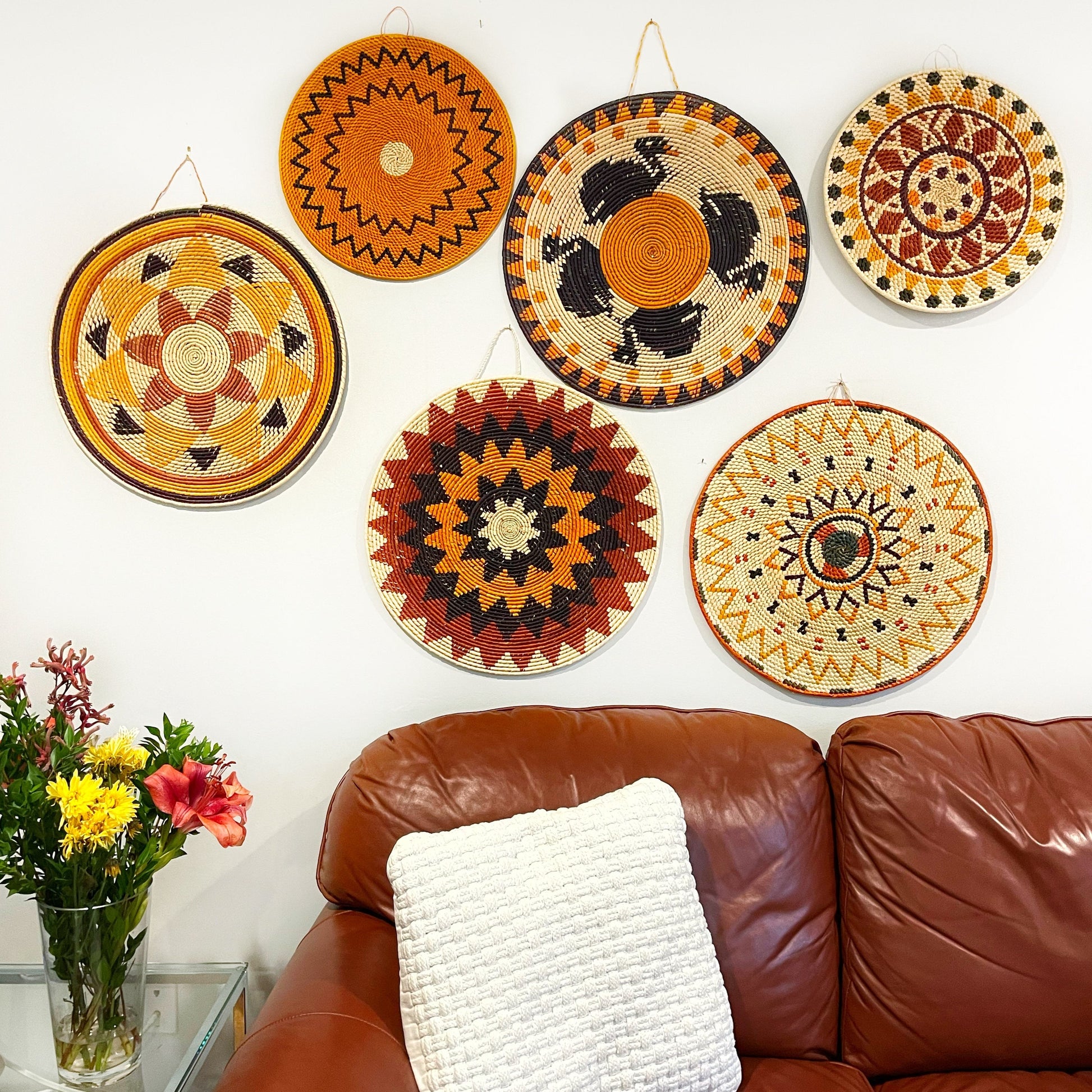 A living room wall is adorned with six colorful Amazonian mandala arts, each with unique patterns of spirals and geometric shapes in shades of red, orange, black, and cream, creating a lively and culturally rich atmosphere.