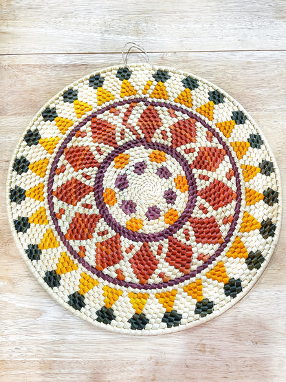 This mandala wall art features intricate weaving in a circular pattern with a mix of cream, orange, burgundy, purple, and dark green, creating a harmonious blend of warm and earthy tones.