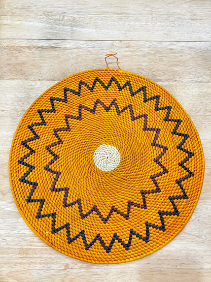 This vibrant mandala wall art showcases a vivid orange backdrop with concentric patterns of jagged black and purple zigzag lines, culminating in a central cream-colored circle. It exudes a lively, energetic aura.