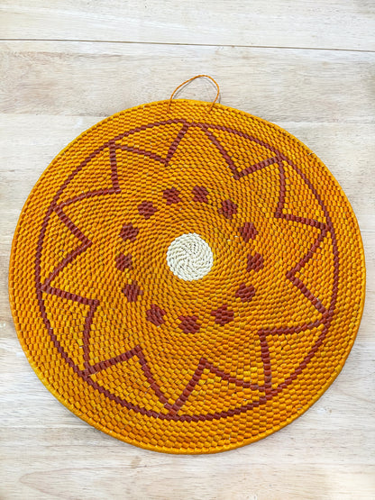 This handwoven mandala features a warm orange base with a symmetrical starburst pattern in shades of rust and brown, converging into a pale cream center. The design radiates outward like a sun.