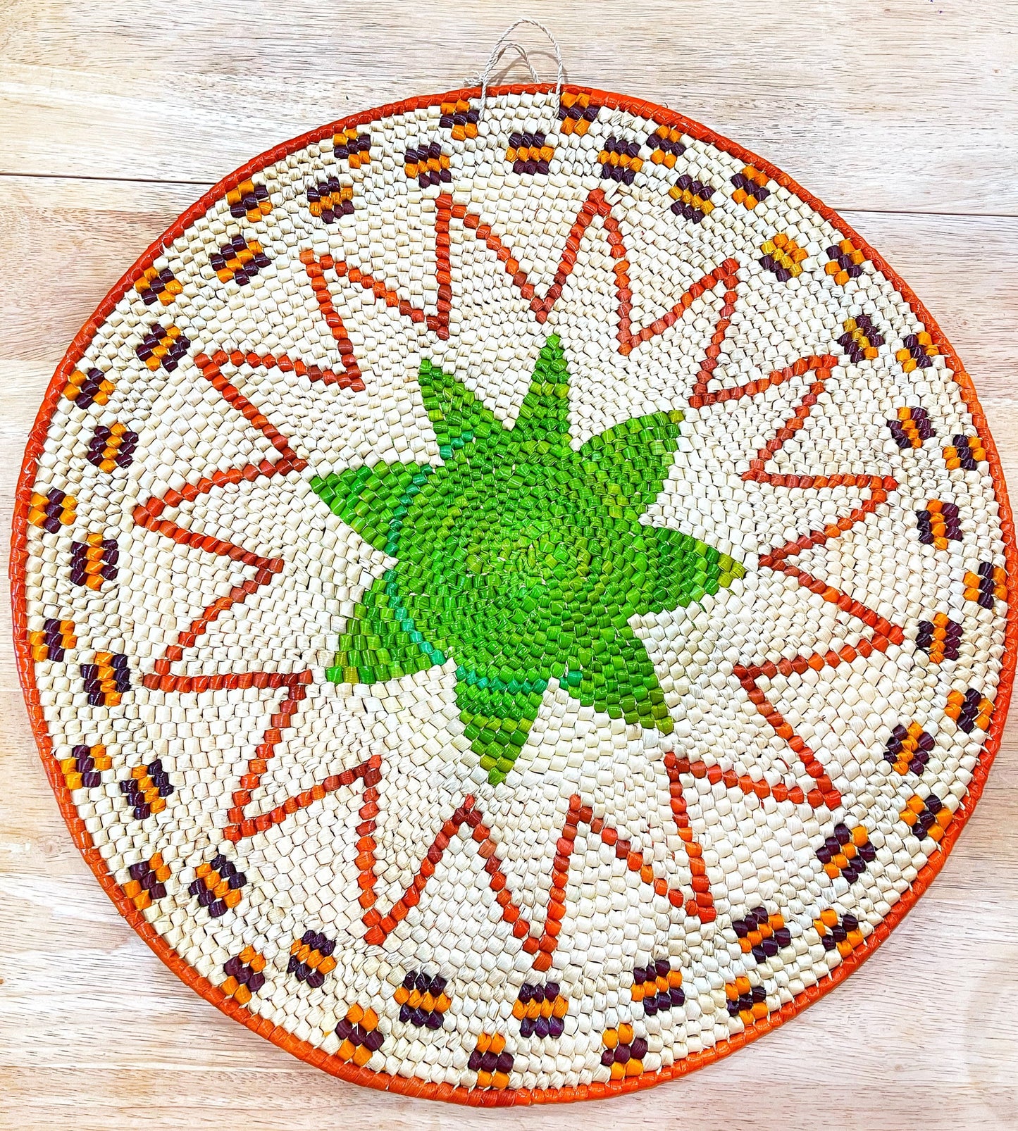 A vibrant, handwoven mandala wall hanging features a green starburst center, surrounded by orange, purple rays and a white, zigzagged border with orange, yellow, and brown accents on a wooden surface.