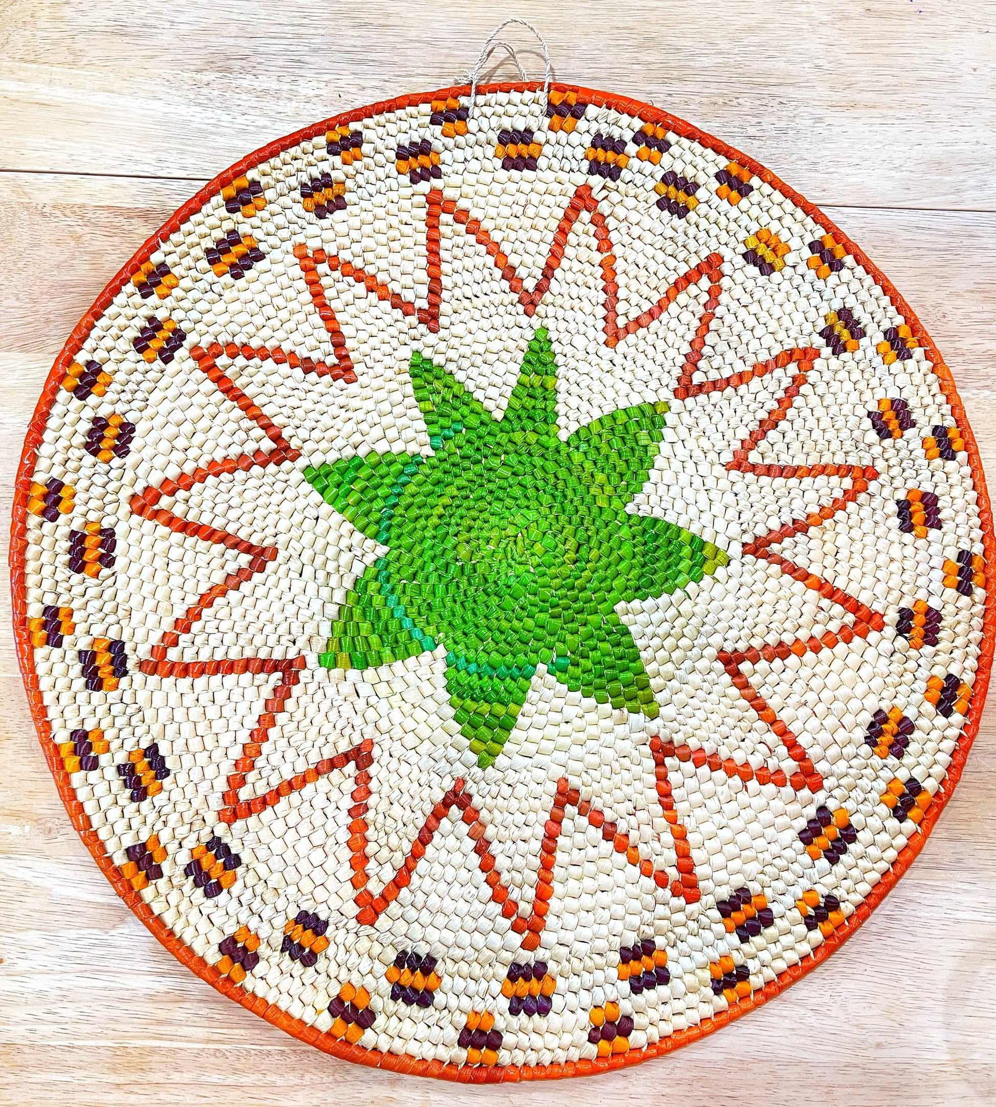 A vibrant, handwoven mandala wall hanging features a green starburst center, surrounded by orange, purple rays and a white, zigzagged border with orange, yellow, and brown accents on a wooden surface.