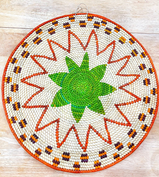 A vibrant, handwoven mandala wall hanging features a green starburst center, surrounded by orange, purple rays and a white, zigzagged border with orange, yellow, and brown accents on a wooden surface.