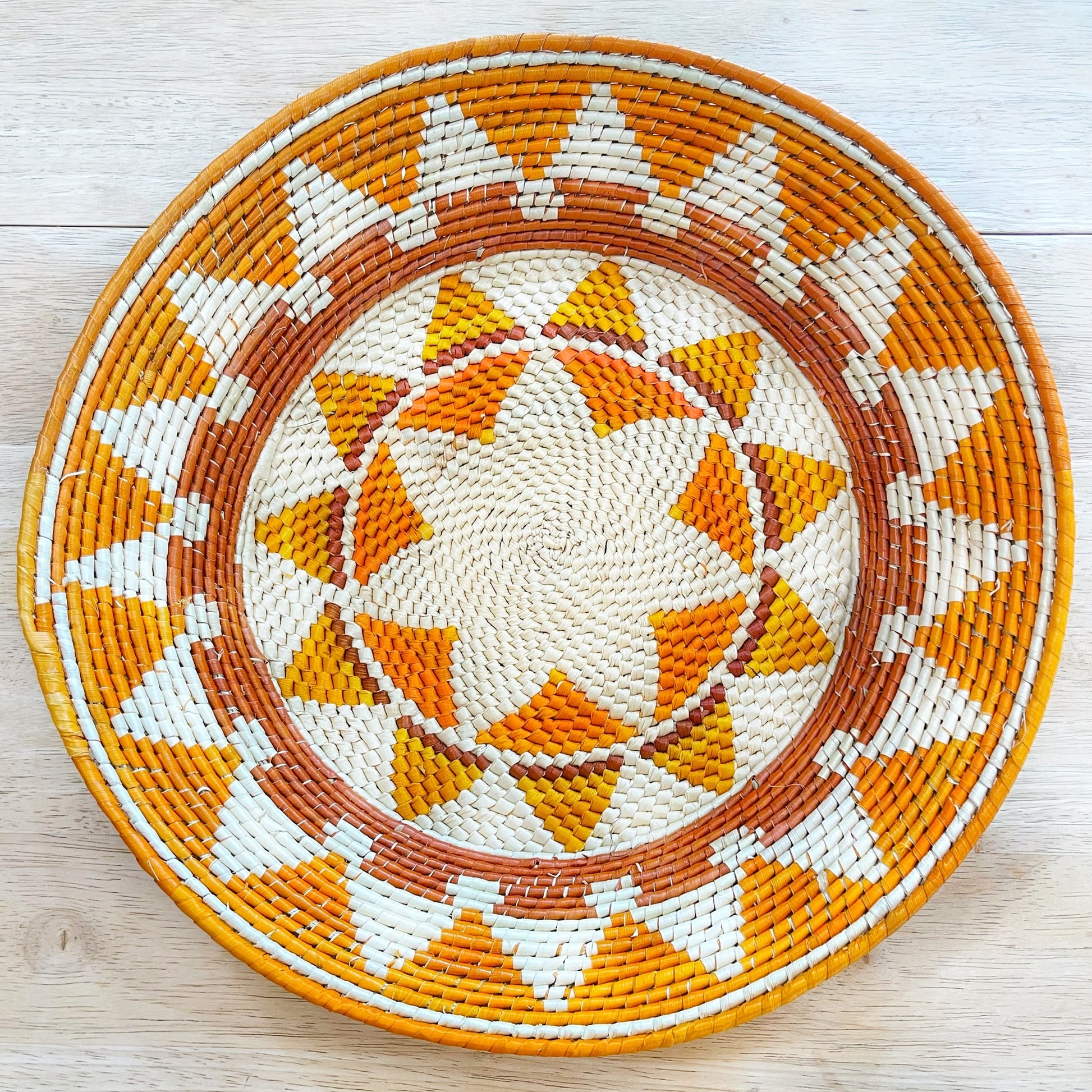 This woven basket displays a vibrant array of colors and patterns. The central design features a complex starburst pattern in shades of beige and vibrant orange. This motif is encircled by a broad border with geometric triangles in white, orange, and a deep brown that gives way to the outermost rim in a solid brown, creating a harmonious blend of warm tones.