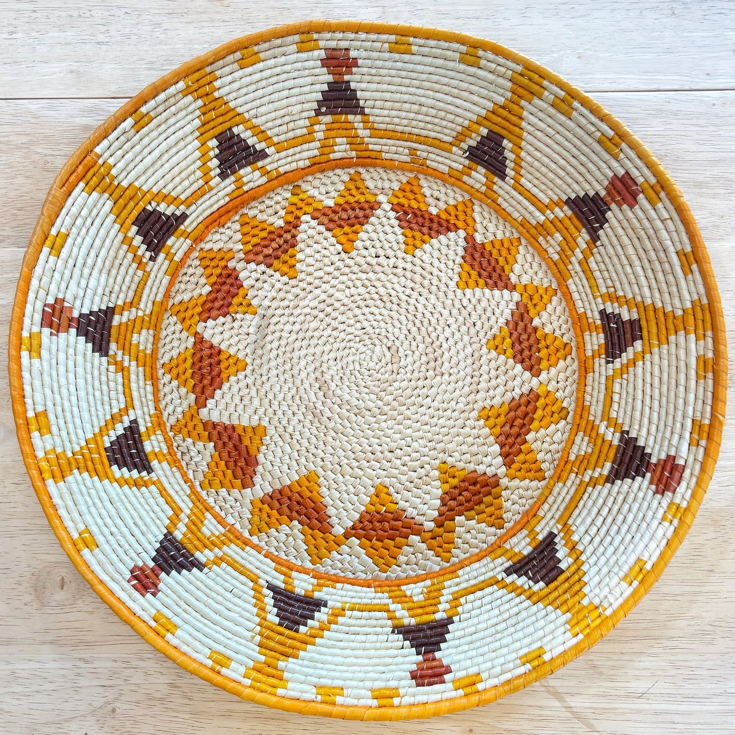 The basket features a central beige area that spirals out from the center, leading to a series of concentric decorative bands. These bands showcase patterns in yellow, white, and dark brown, with the addition of a vibrant orange creating a star or sunburst design. The outermost ring is a rich yellow, framing the intricate design work within.