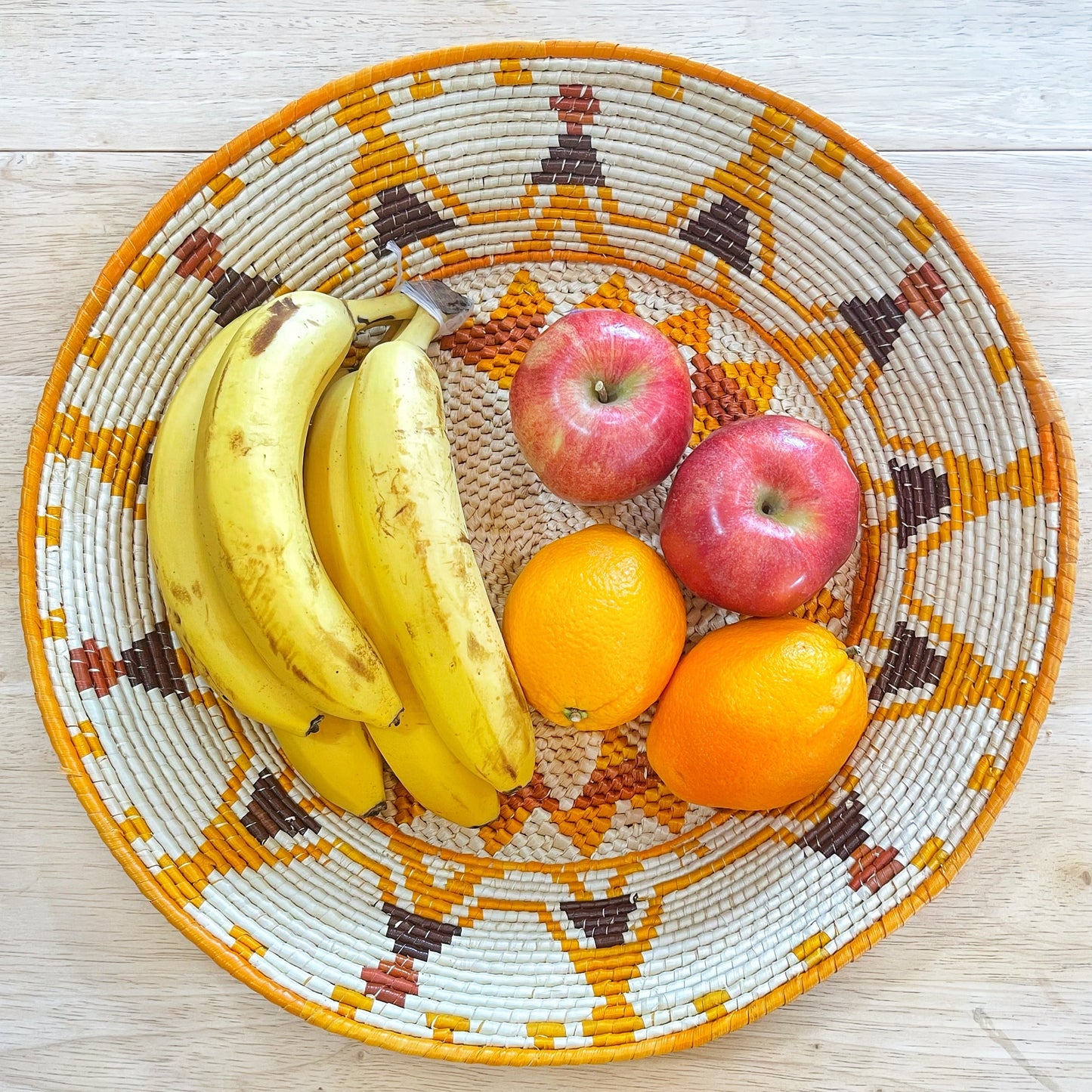 The basket is filled with ripe bananas, which are bright yellow, alongside vivid orange oranges and red apples, all resting atop the intricate pattern of the woven surface. The mix of fruits adds a lively and colorful touch to the detailed craftsmanship of the basket.