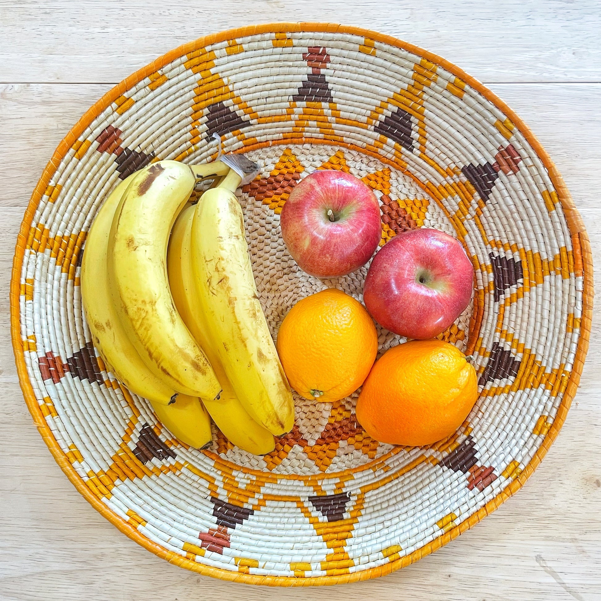 The basket is filled with ripe bananas, which are bright yellow, alongside vivid orange oranges and red apples, all resting atop the intricate pattern of the woven surface. The mix of fruits adds a lively and colorful touch to the detailed craftsmanship of the basket.