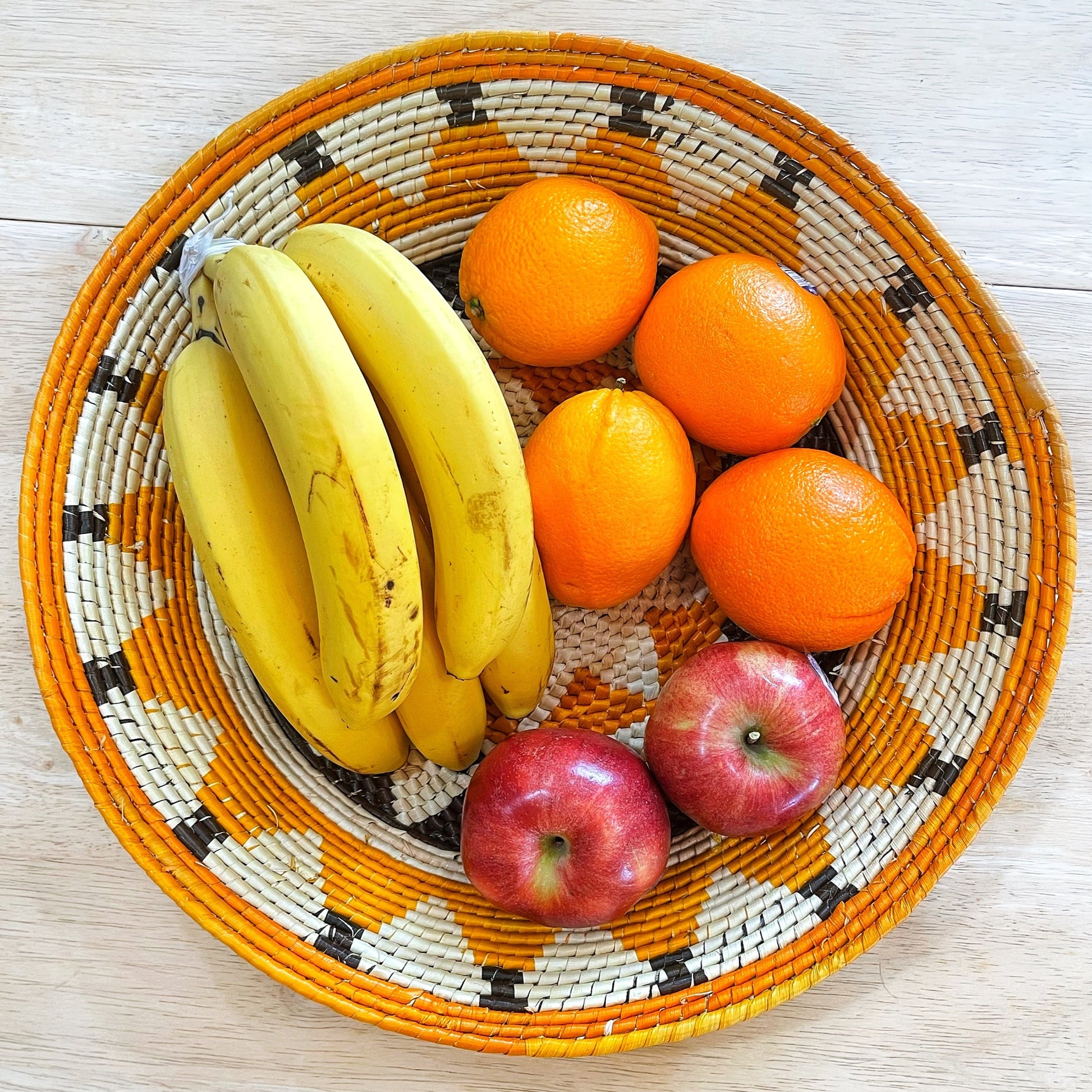 The basket brims with a collection of fresh fruits: a cluster of yellow bananas nestles beside vibrant orange oranges and two glossy red apples, all set against the basket's handwoven backdrop with orange and black geometric patterns and a beige central area.