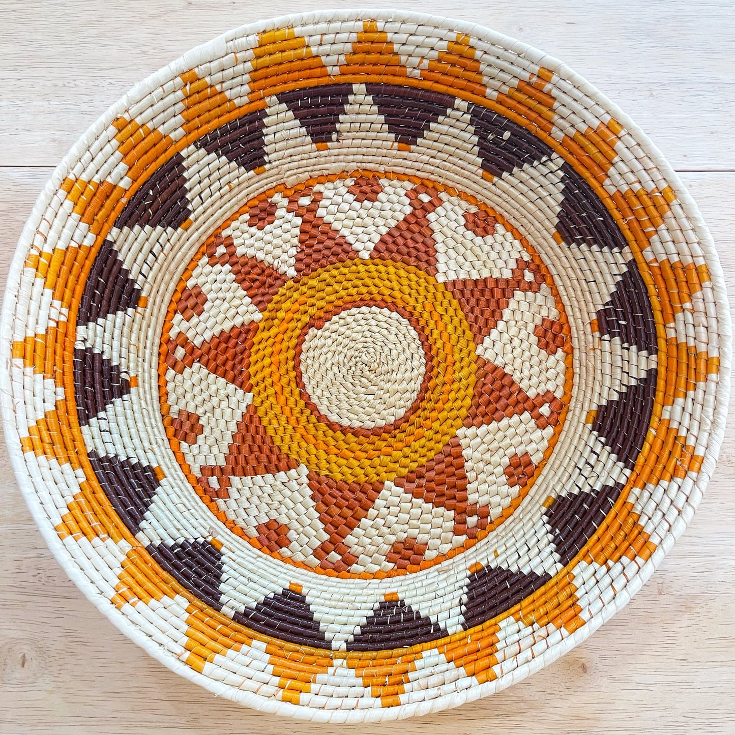 The image showcases a handcrafted basket with an elaborate design woven in natural beige, deep brown, and bright orange fibers, creating a concentric circular pattern reminiscent of a sunburst. The outer rim is a thick band of beige, framing the detailed interior pattern.