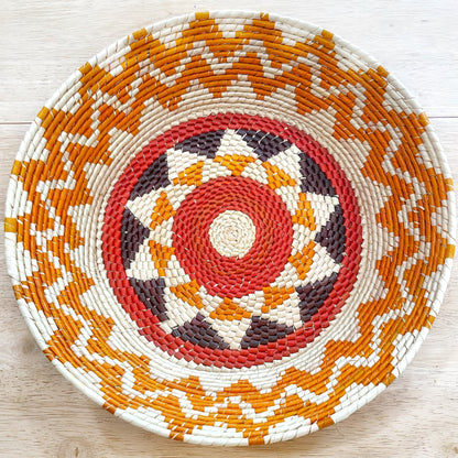 The basket displays a striking pattern starting from a central beige circle, expanding into a vibrant red ring, followed by rings in white, brown, orange, and red, creating a bullseye effect. The outer sections exhibit a checkerboard-like pattern in orange, white, and brown, encased by a thick beige border.