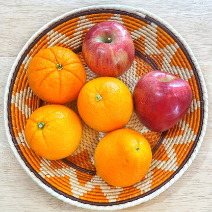 A handwoven Amazonian bowl crafted from tucuma palm fibers filled with vibrant fruits. The bowl features natural beige tones with striking orange and brown geometric patterns along the rim, cradling bright oranges and a glossy red apple, illustrating its utility and size