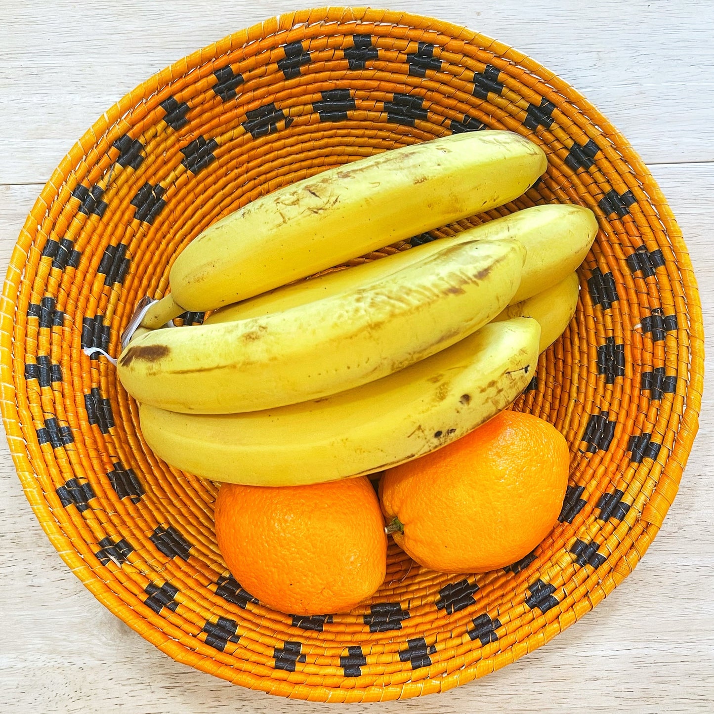 A vibrant handwoven bowl made from tucuma palm fibers, filled with fresh bananas and oranges. The bowl features a striking pattern of orange and black squares on the rim, perfect for fruit display or decoration