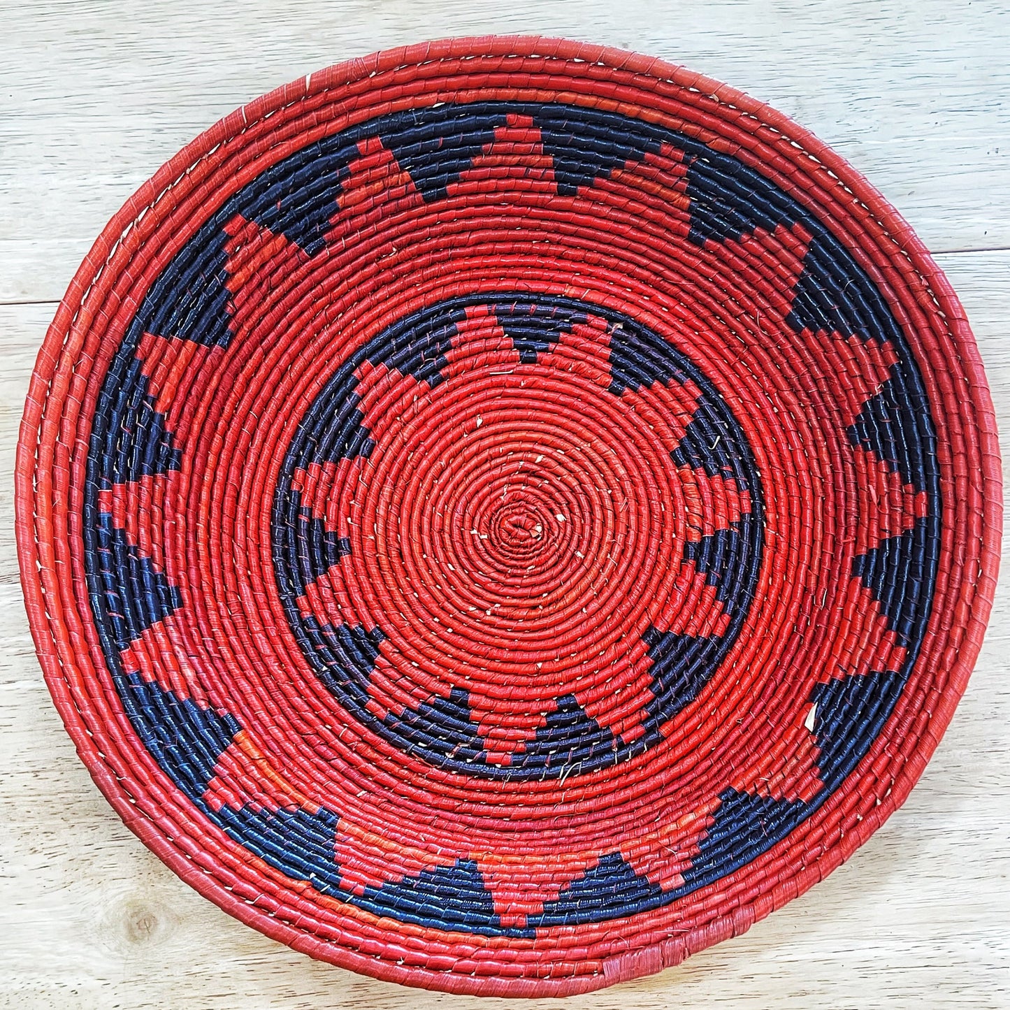 The bowl features a bold and intricate pattern with a striking color palette. The central spiral is a rich red, which expands outward in alternating bands of red and blue. The pattern transitions into a sunburst design, with blue spikes against the red background, enclosed by a broad red rim with a black zigzag pattern.