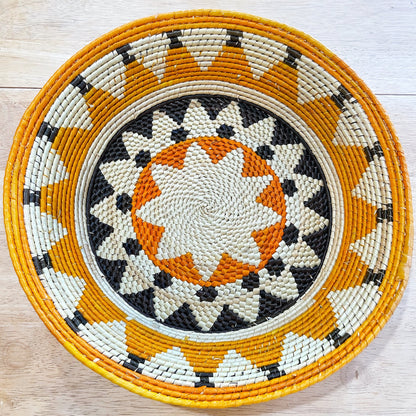 The image displays a handwoven basket with a captivating starburst pattern in the center, featuring a mix of natural beige, bold black, and bright orange fibers. Surrounding the starburst is a ring pattern in beige, black, and orange, followed by an outermost rim in a sunny orange hue. The design showcases the intricate artistry of the weave.