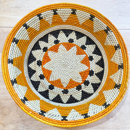 The image showcases a handwoven bowl with a stunning geometric pattern. The central motif is a starburst design in shades of beige and orange, surrounded by black accents that create a dynamic contrast. This is encircled by a series of concentric rings alternating in colors of beige, orange, and black, with the outermost ring in a bright orange, giving the bowl a vibrant and eye-catching edge.