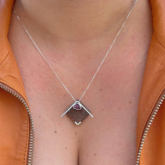 A 925 sterling silver pendant with an amethyst gemstone and a base of recycled materials including açaí and peach palm rests gracefully on a silver chain against a person's neck, framed by an orange jacket.