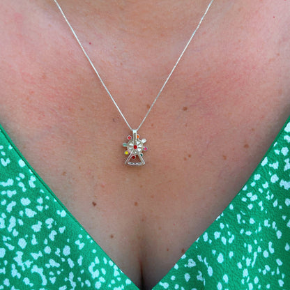 A silver pendant  shaped like a mini Ferris wheel, with colorful inlays hangs on a delicate chain against skin, paired with a green patterned garment, showcasing artisanal charm.