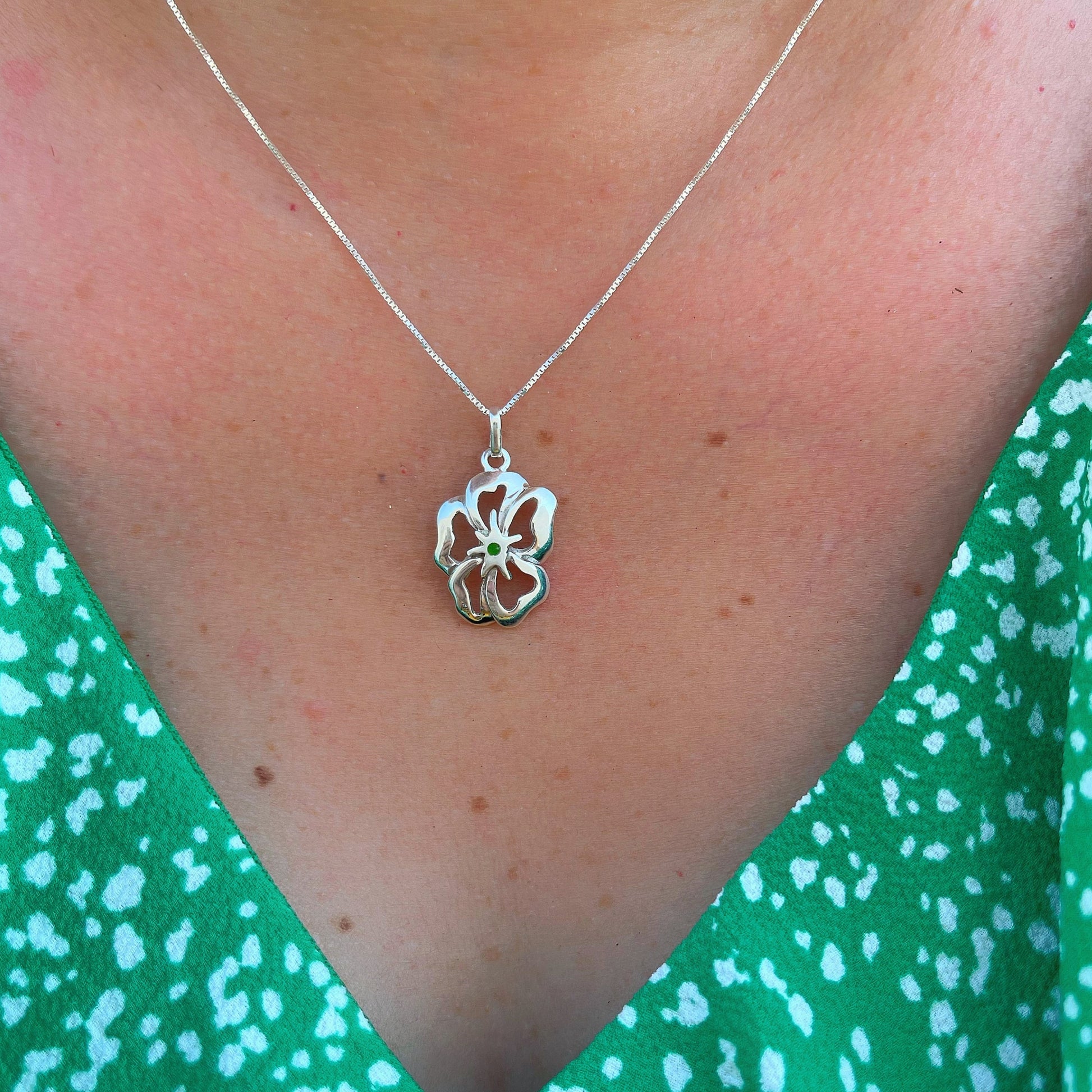 A silver flower pendant with a green malachite inlay on a necklace, set against a green patterned fabric, evoking a fresh, natural aesthetic.