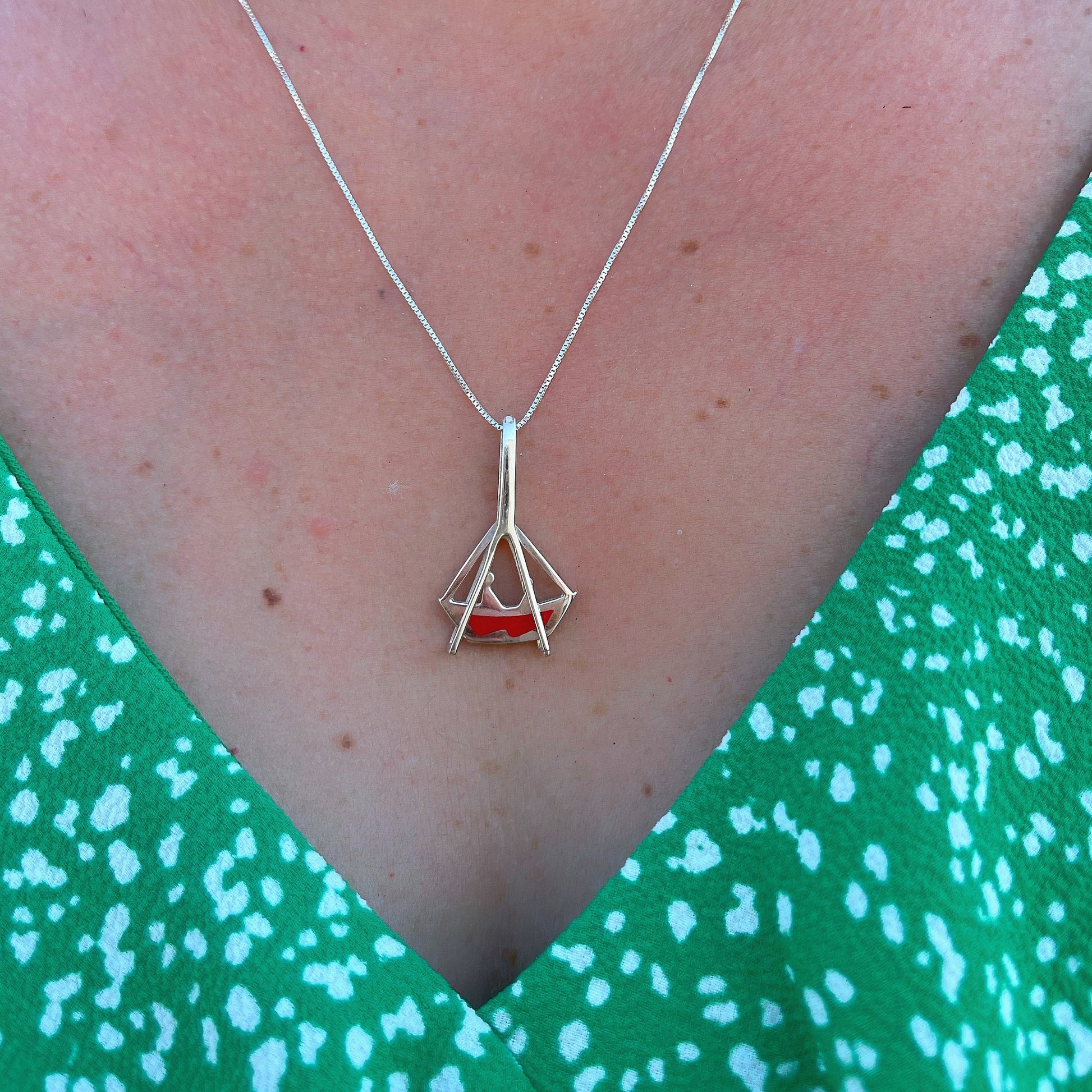  A sterling silver necklace with a unique boat-shaped pendant resting above a green patterned garment. The pendant has a red inlay, reflecting the traditional craft techniques of Belém, Brazil, and is a homage to the city's festive religious celebrations.