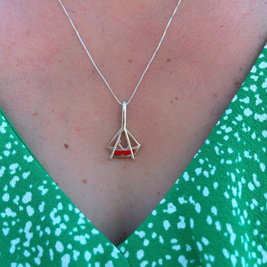  A sterling silver necklace with a unique boat-shaped pendant resting above a green patterned garment. The pendant has a red inlay, reflecting the traditional craft techniques of Belém, Brazil, and is a homage to the city's festive religious celebrations.