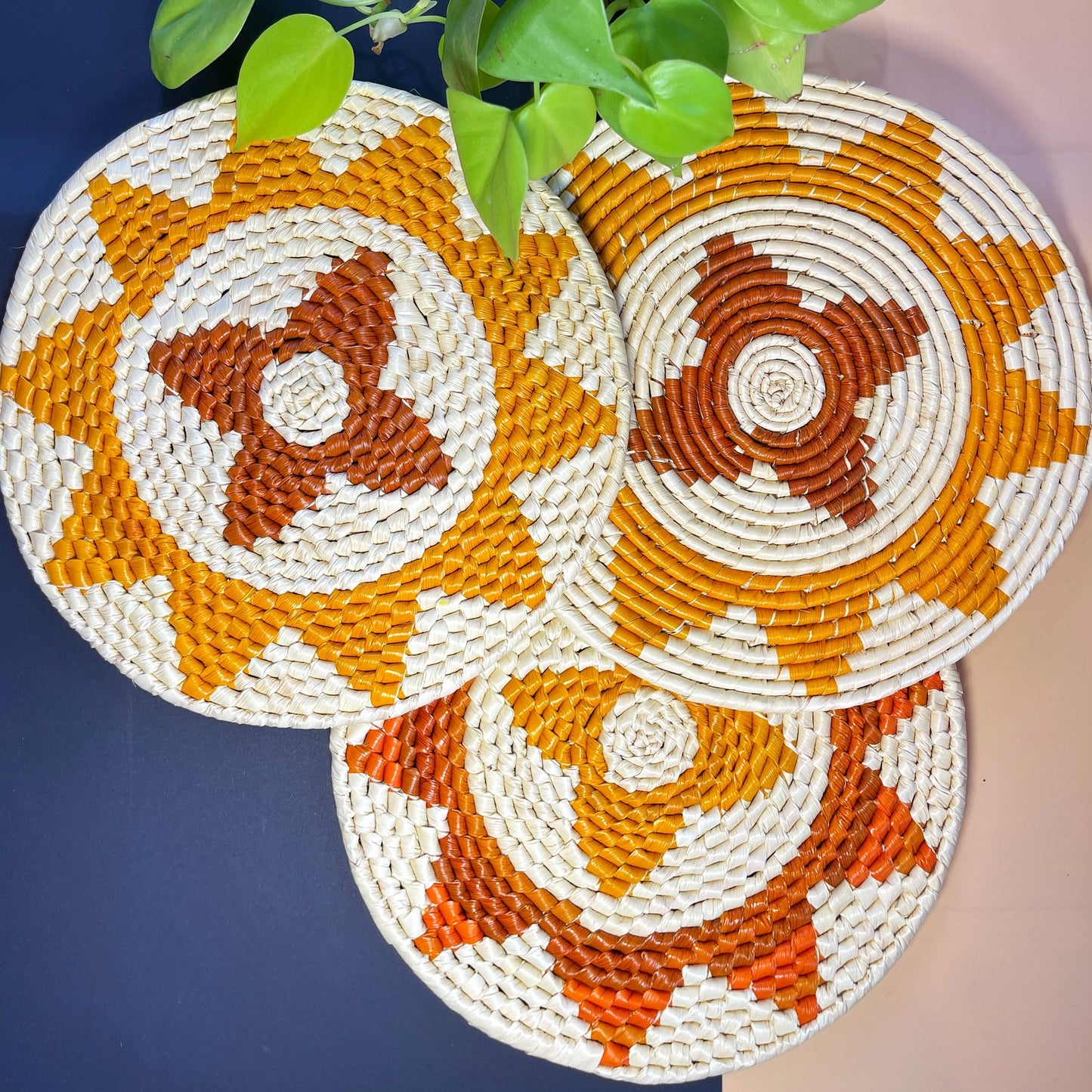 Three circular woven trivets with white, orange, and brown spiral patterns, beside green leaves on a navy surface.