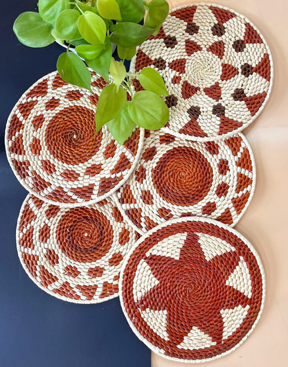 Five handcrafted trivets arrayed on a table, woven from natural fibers into intricate patterns with a rich terracotta, deep brown, and cream color scheme, accompanied by a sprightly green plant.