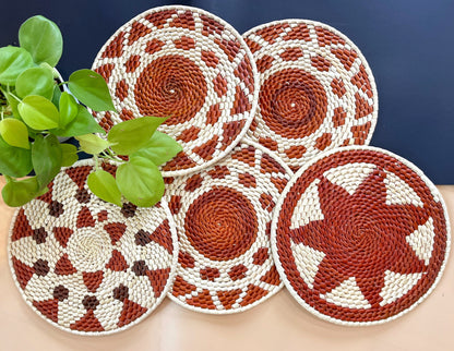 Five handwoven trivets in terracotta, brown, & cream. Intricate patterns adorn the natural fibers.