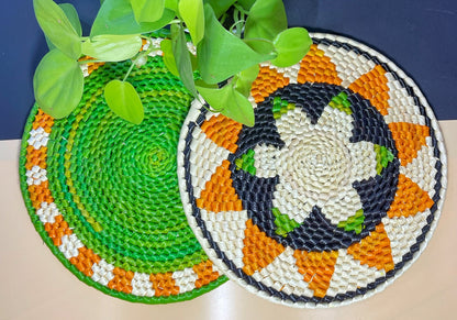 The image showcases two vibrant handwoven trivets. One features a bright green center, beige and orange border. The other boasts beige, green, and dark blue with a patterned black, orange, and green border. A lush green plant adds a natural touch.