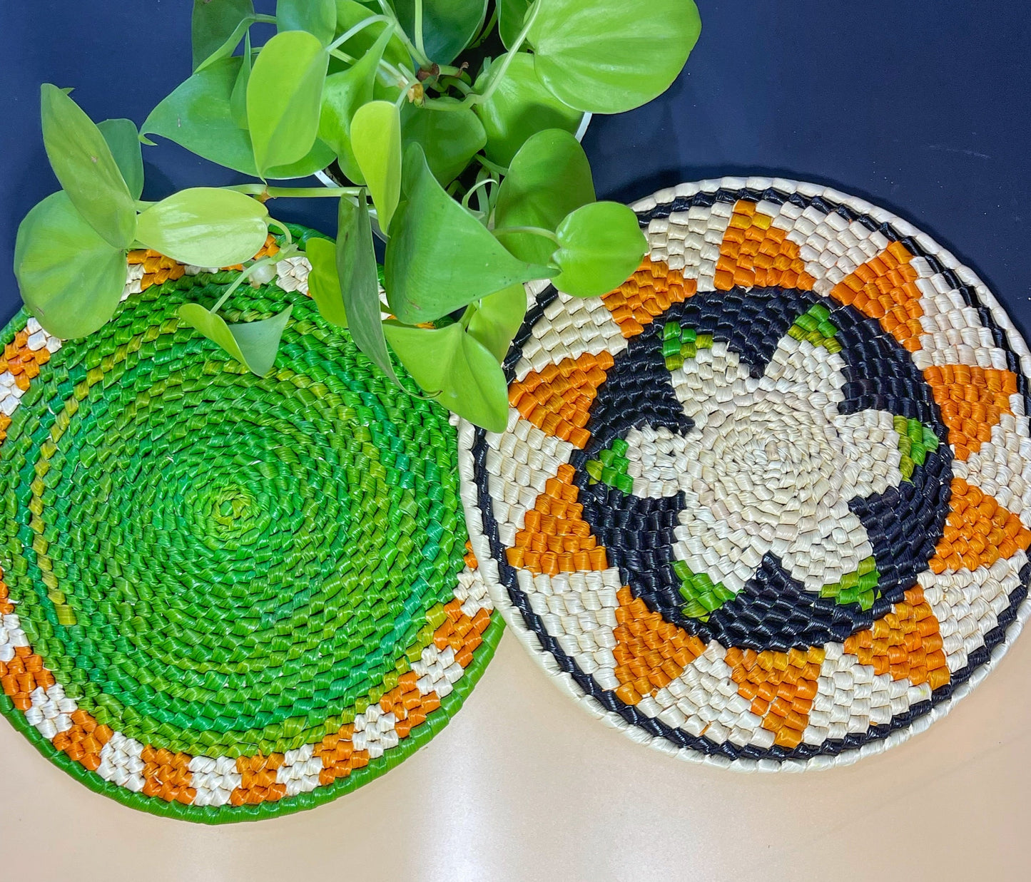 Two colorful handwoven trivets: one with a green center & beige/orange border. The other features beige, green & dark blue with a black, orange & green patterned border.