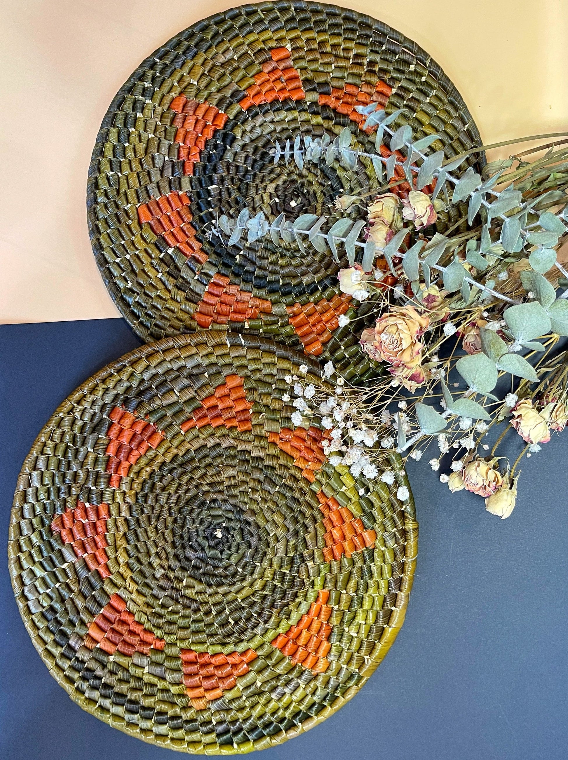 Two handwoven trivets display a striking pattern with olive green bases and concentric circles in shades of green, accented with pops of burnt orange, alongside dried floral arrangements.