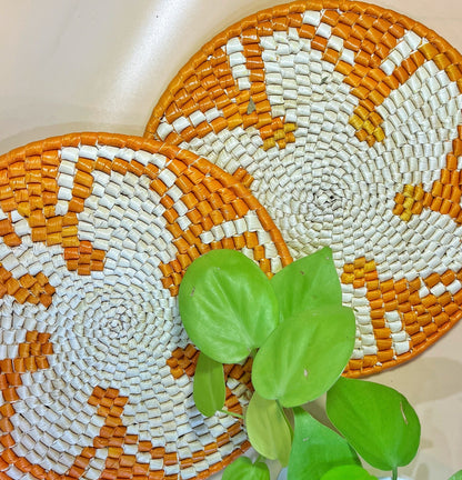 The image features two handwoven trivets with a white base and intricate patterns in shades of orange, adorned with a green plant adding a lively contrast.