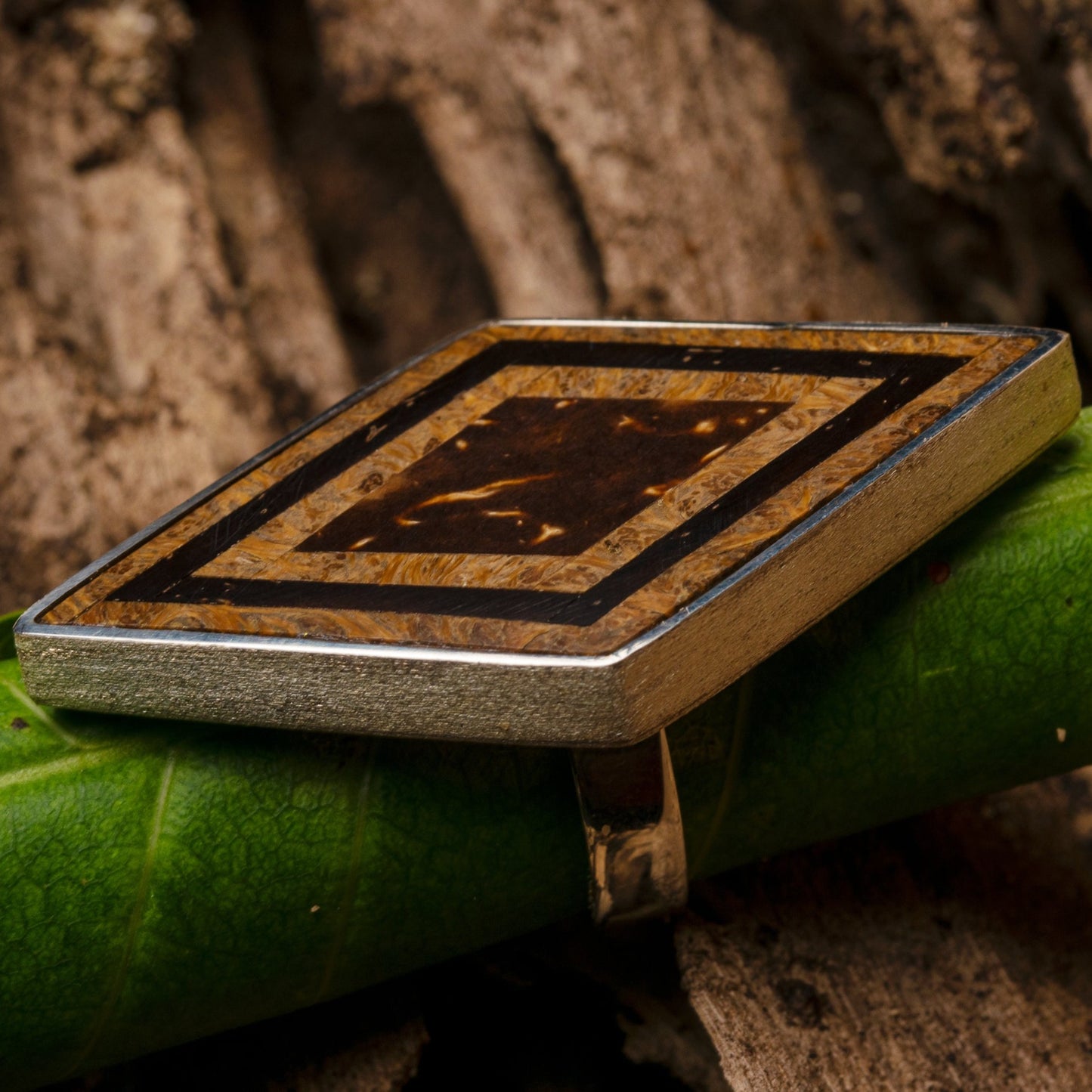 A close-up of an elongated hexagonal .925 sterling silver ring with a marquetry top of babassu, Brazil nut husk, and coconut, resting on a green leaf.