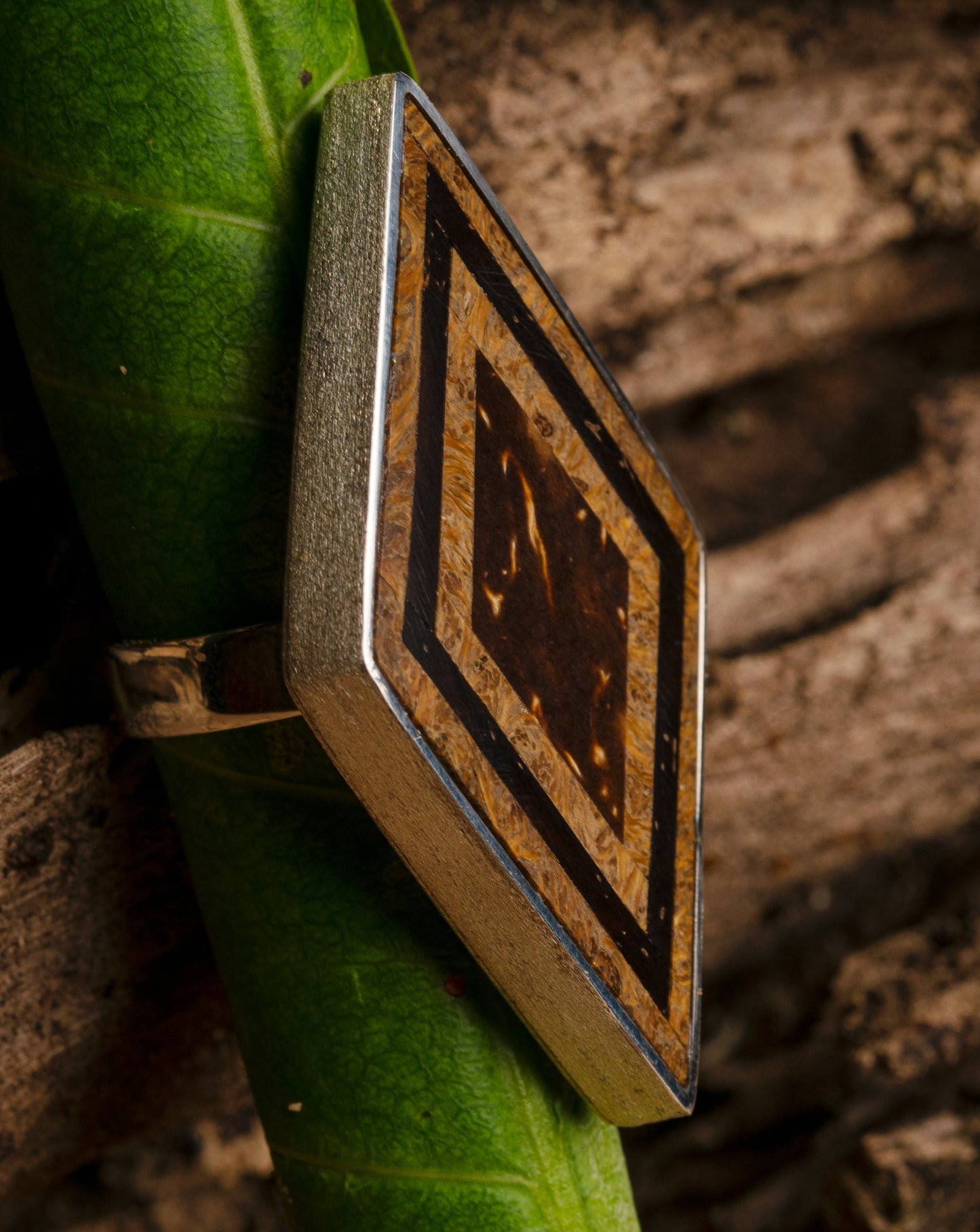 The ring, with its striking seed marquetry, is displayed against a natural backdrop of a green leaf and rough, textured wood, emphasizing its organic beauty. 