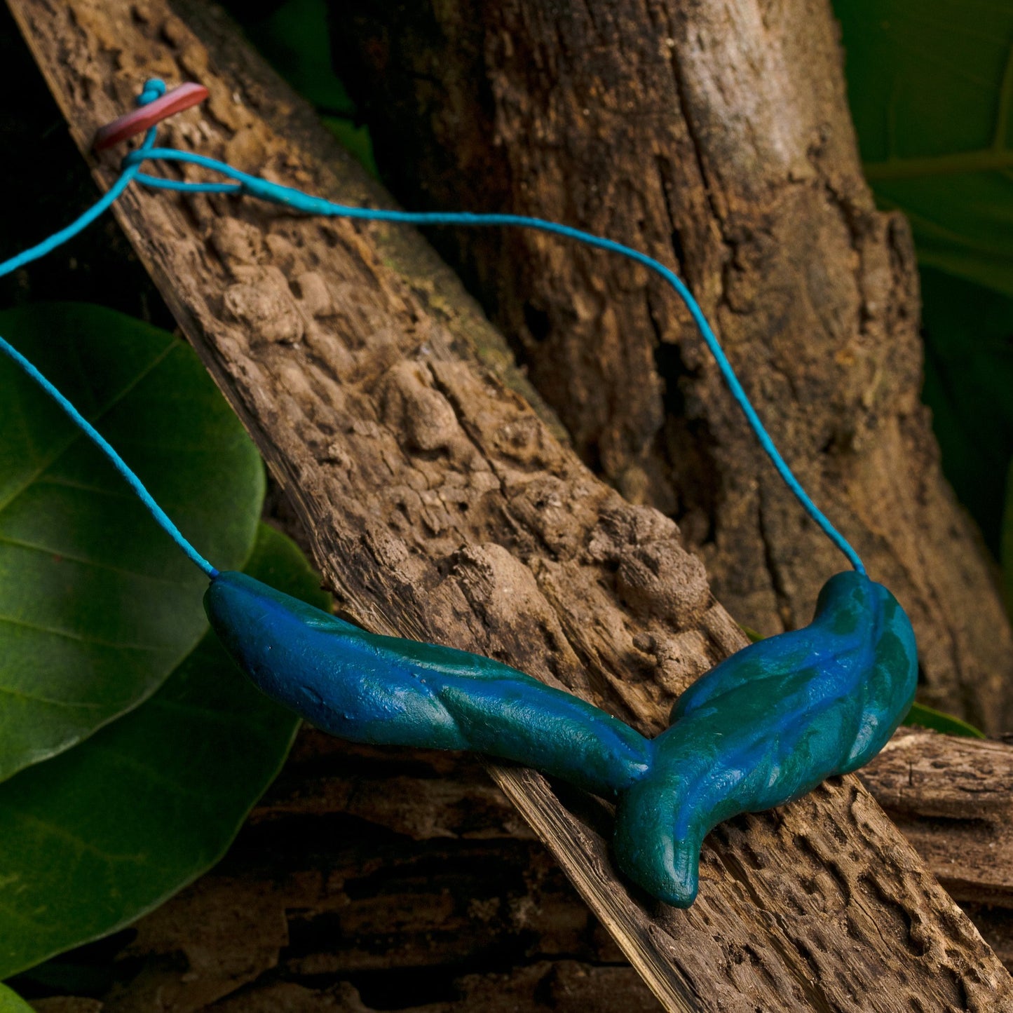 Artistic blue rubber necklace on a bright açaí fiber cord, set naturally on textured wood among green leaves, showcasing handcrafted beauty