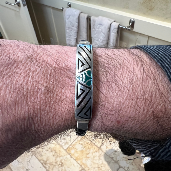 A bracelet on a wrist showcasing a silver band with tribal patterns, a central inlaid turquoise muiraquita—the Amazonian frog symbol for protection—and additional design details.