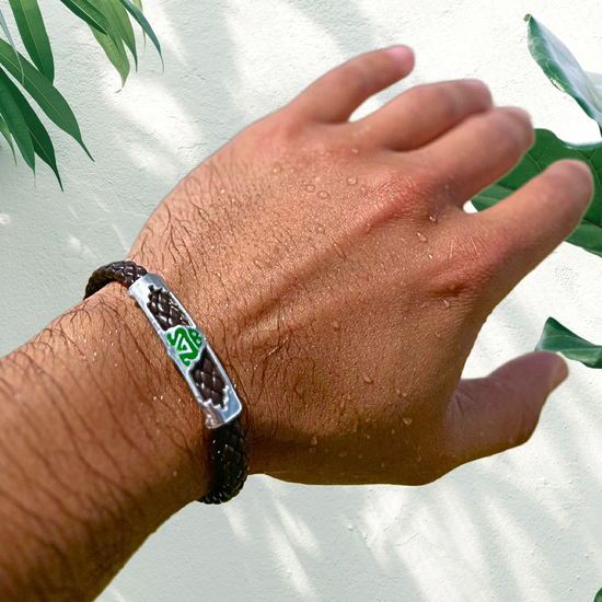 The bracelet on the person's wrist is handcrafted, incorporating vegan leather for the dark brown braided straps, and sterling silver for the silver tag in the center. The green and white emblem is a muiraquitã, a traditional Brazilian amulet said to bring good luck, which adds cultural significance and a pop of color to the piece. The combination of materials and the inclusion of the muiraquitã make this bracelet both stylish and meaningful