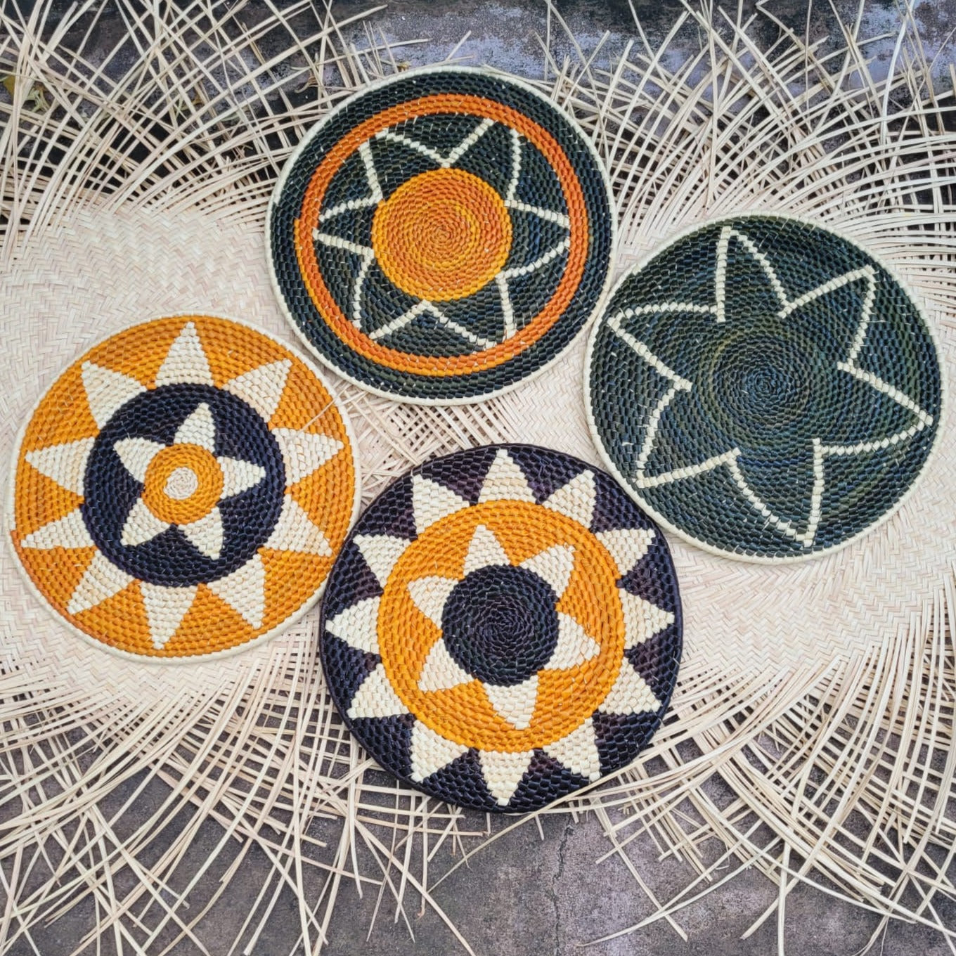 Four handwoven tucuma placemats displayed on a woven background. Each features starburst patterns in colors of navy, green, orange, and white, with one showcasing a green star on navy.