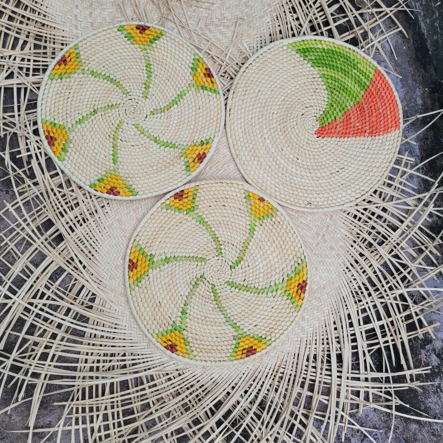 Three round tucuma fiber placemats with pinwheel and avocado designs in cream, green, yellow, and red rest on a backdrop of loose palm strands.