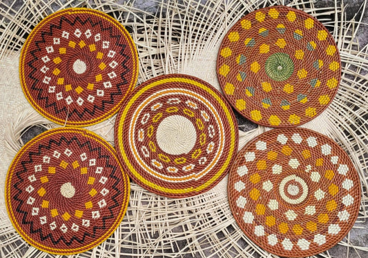 This image shows a collection of five round, hand-woven placemats with intricate patterns, displayed on a wicker surface. The placemats feature warm earthy tones—reds, yellows, browns, and natural hues—in a variety of designs, including concentric circles and geometric shapes.