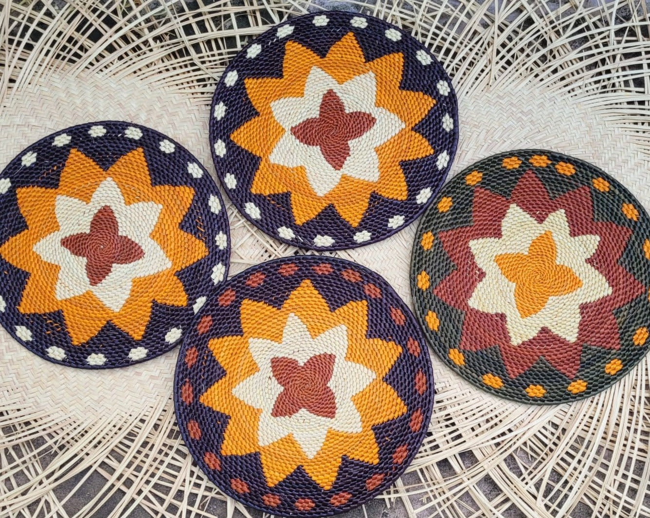 Four round tucuma placemats with central floral motifs in navy, orange, white, and burgundy, on a woven backdrop.