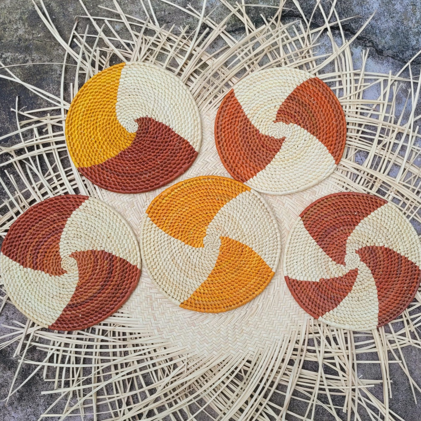 Five cream-colored handwoven trivets, each with a swirl design in alternating shades of orange and brown, placed on a herringbone pattern wicker background.