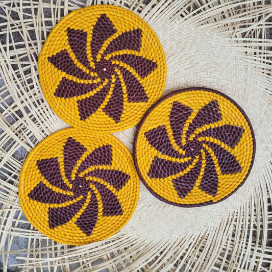 Three round handwoven trivets with a yellow base and dark purple spiral patterns, made from tucuma palm fibers, on a wicker background.